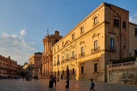 Large building with classical architecture on a sunny square with passers-by, Ortigia Island, Isola