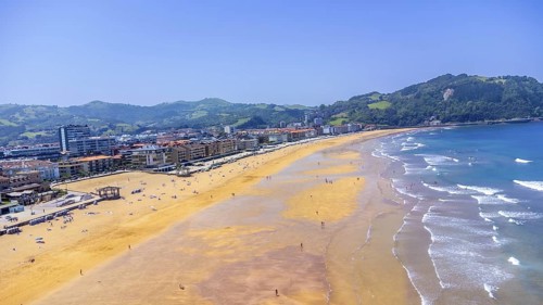 Aerial view of zarautz beach during a sunny summer day, featuring golden sand, blue water, and lush