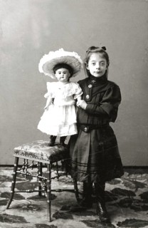 Girl doll dresses, 1930s, girl standing next to a chair carrying a doll in a dress, both in