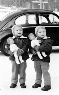 Twins, 1950s, Two children standing outside in the snow, holding dolls, car in the background,
