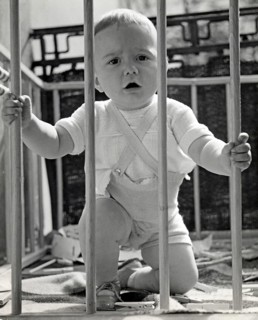Little boy in a playpen, 1930s, Toddler kneeling in a cot and holding on to the wooden railing,
