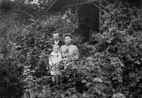 Family standing at a wooden hut, 1920s, adults with child standing in front of a house surrounded