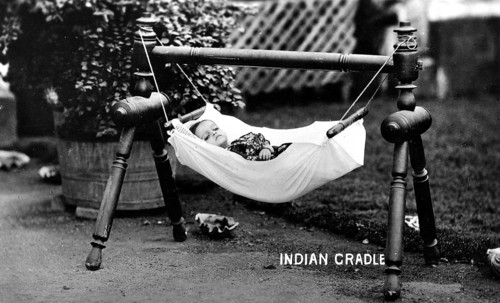 Baby hammock cradle, 1930s, A baby lies in a hanging cradle in the garden, surrounded by plants,