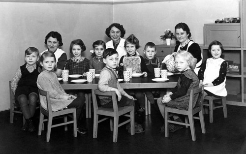 Kindergarten group, 1930s, Kindergarten scene with seated children, woman at the table with cups,