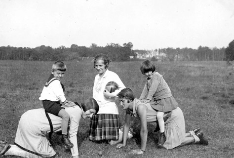 Two children riding on the backs of their mothers, 1930s, Children playing in the meadow with two