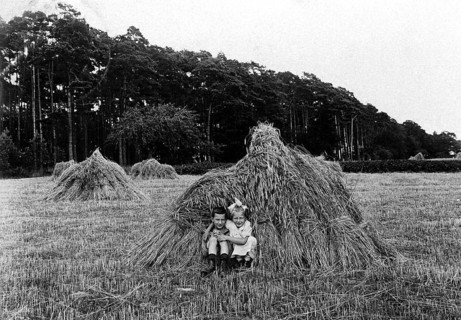 2 children in a haystack, 1920, Two children sitting in a field in front of a large bale of straw,