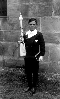 Boy has communion, 1920s, A boy stands with a communion candle and a book in front of a stone wall,