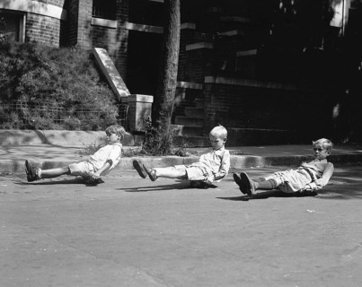 Three children sitting on skateboards, 1920s, Three boys playing on skateboards on a city street,
