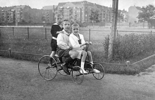 Three children on a historical bicycle, 1910s, Three children riding a tricycle on a cobbled