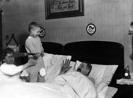 Children teasing father, 1920s, Children playing with a man in bed while one smiles and the other