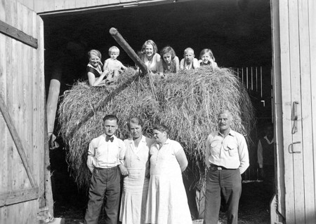 Farming family in front of the barn, ca. 1930, Family posing smiling in front of a bale of hay in