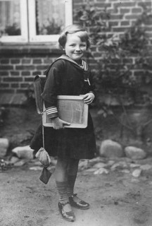 School enrolment, 1920s, A girl with a slate and school uniform stands in front of a brick wall,