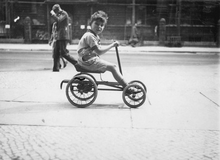 Little boy on a quadricycle, 1920s, Boy on a tricycle in an urban environment with passers-by in