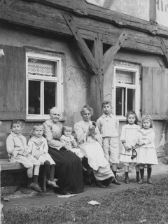 Two old ladies with children, 1920s, A large family sits and stands on a bench in front of a rustic