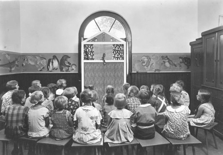 Puppet theatre, 1920s, children sit in a classroom and watch a puppet show. The walls are
