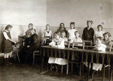 Sewing class at school, 1920, girls in a classroom practising sewing while a teacher sits at a
