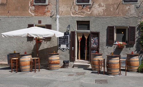 Enoteca with wooden barrels as tables and bar stools under a large parasol in front of an old house