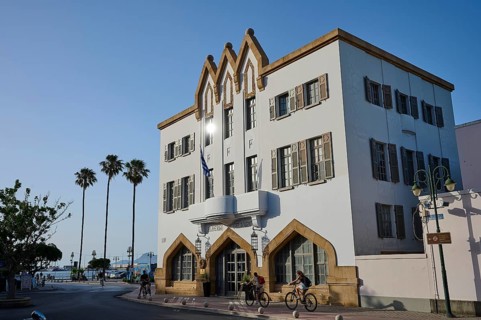 Imposing building in a Mediterranean setting with palm trees and cyclists under a bright blue sky,