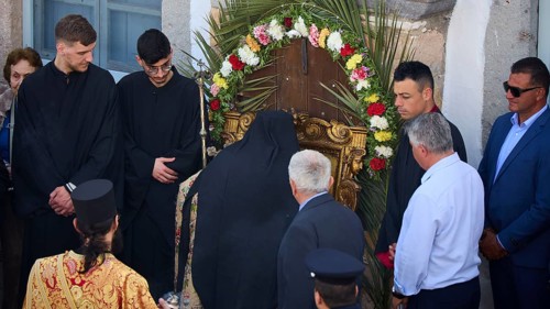 A priest prays in front of a wreath decorated with flowers, surrounded by people in traditional and