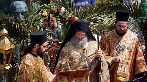 Priests in richly decorated robes reading from a book during a solemn ceremony under palm trees,