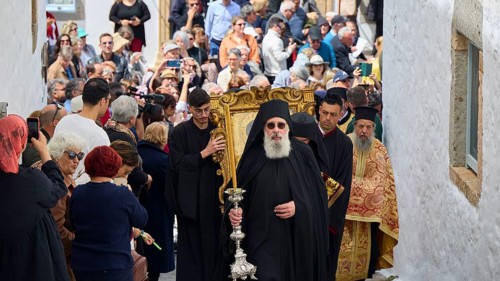 A priest leads a procession through a narrow street, followed by worshippers and surrounded by a