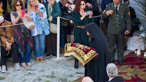 A priest in a black cloak with golden embroidery at a religious event, surrounded by people taking