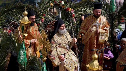 Priests in elaborate robes sit surrounded by palm trees during an outdoor religious ceremony,