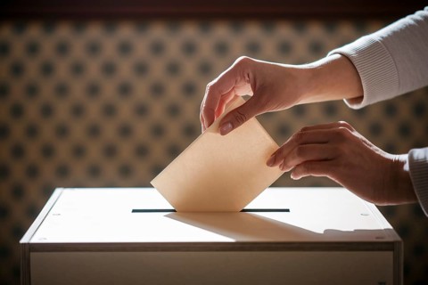 Woman's hands casting vote by placing folded ballot paper into voting box during election.