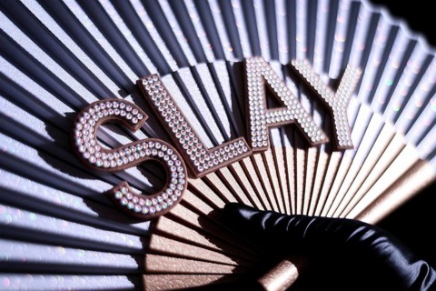 Close-up of drag queen's rhinestone SLAY fan held in black satin glove with glitter and warm