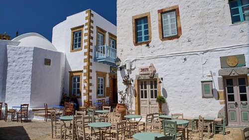An outdoor area of a café with empty tables and chairs in front of white buildings on a sunny day,