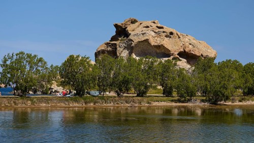 Rock rises behind a row of trees on the lakeshore under a clear sky, Rock of Kalikatsou, Seasonal