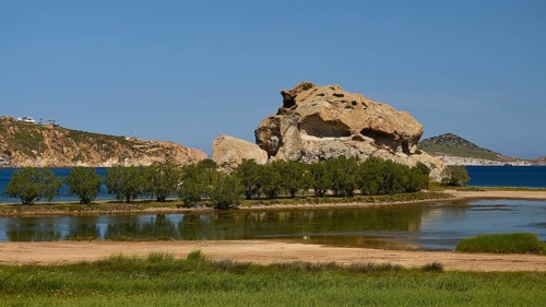 Massive rock formation surrounded by water and trees under a clear sky, seasonal salt lake, Grikos,