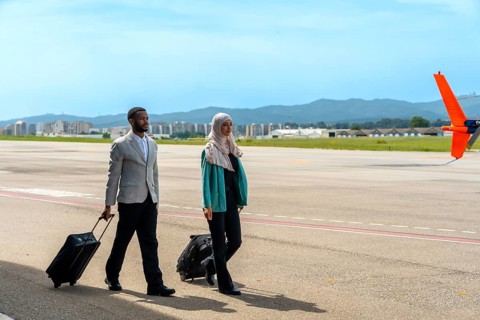 Businessman and businesswoman walking along the airfield tarmac, pulling carry on luggage while
