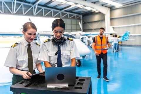 Two female colleagues pilots preparing flight plan using laptop and writing on clipboard in hangar