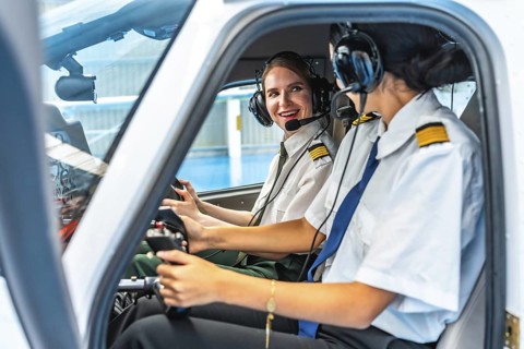 Two smiling female pilots wearing headsets and uniforms are sitting in the cockpit of a small