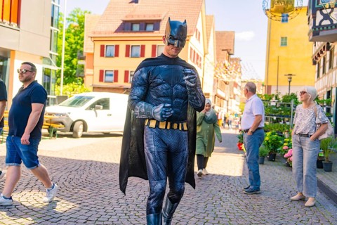 Man in Batman costume walking in a busy sunny city centre, Calw train station festival, Black