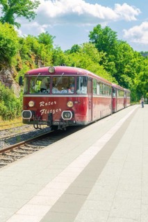 Red train stops in a rural setting on a sunny platform under a blue sky, Calw railway station