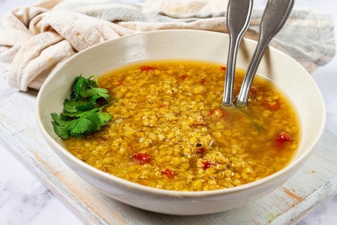 A warm lentil soup with a rich golden color is served in a rustic bowl. The soup features fresh
