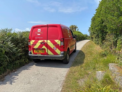 A red Royal Mail van on a rural road surrounded by lush greenery under a sunny sky, St Agnes, Isles