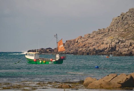Small boat with orange sail in calm coastal waters near rocky cliff, Tresco, Isles of Scilly,