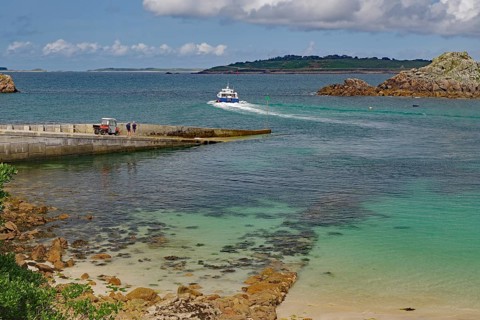 A boat approaches the harbour on clear blue water surrounded by rocks and islands, Ferry, St Agnes,
