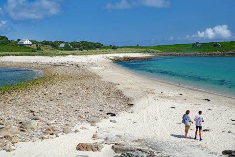 Two people walking along a soft sandy beach with blue sea and sky, St Agnes, Isles of Scilly,