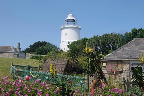 A lighthouse next to a sunny garden with blooming flowers and stone houses under a blue sky, St