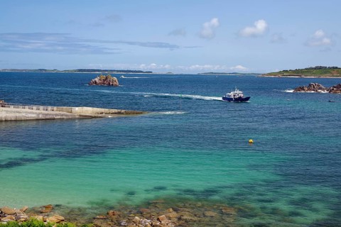 A boat sails on the open sea between small islands, under a clear blue sky, St Agnes, Isles of