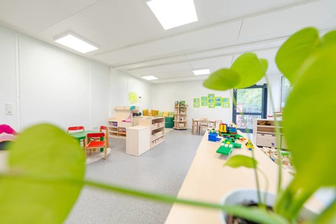 Bright classroom with play tables and a large plant in the foreground, transitional containerised