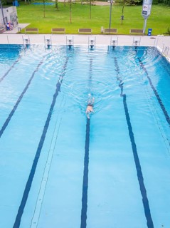 A person in a turquoise-coloured swimming pool swims laps surrounded by empty lanes on a warm