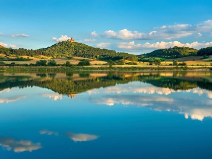 Wachsenburg Castle is reflected in the still water of a lake under a blue sky with cumulus clouds,