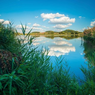 Wachsenburg Castle is reflected in the still water of a lake under a blue sky with cumulus clouds,