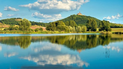 Wachsenburg Castle is reflected in the still water of a lake under a blue sky with cumulus clouds,
