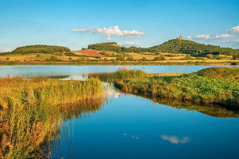 Wachsenburg Castle is reflected in the still water of a lake under a blue sky with cumulus clouds,